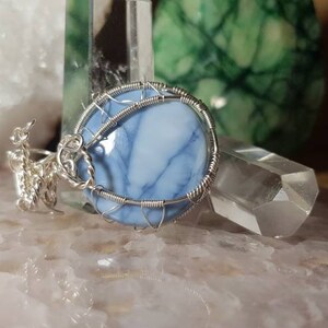 May include: A silver wire wrapped pendant featuring a blue and white opal stone. The pendant is displayed on a white surface with other crystals.