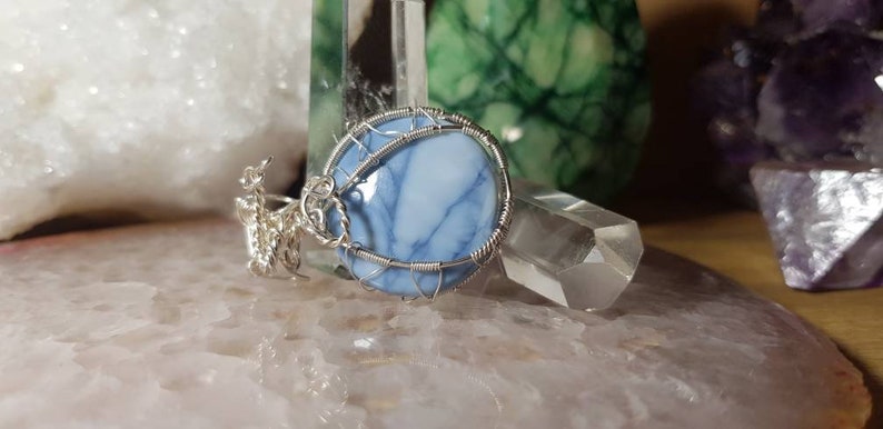May include: A silver wire wrapped pendant featuring a blue and white opal stone. The pendant is displayed on a white surface with other crystals.