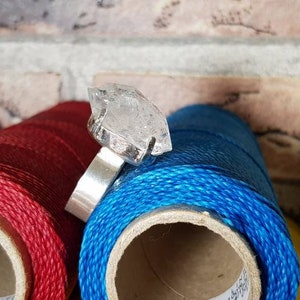 May include: A silver ring with a clear quartz crystal set in the center. The ring is sitting on top of spools of red, purple, and blue thread.