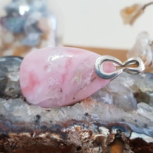May include: A pink and white gemstone pendant with an infinity symbol charm. The pendant is set in silver and is displayed on a rock.