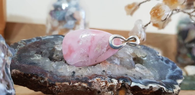 May include: A pink and white gemstone pendant with an infinity symbol charm. The pendant is set in silver and is displayed on a rock.