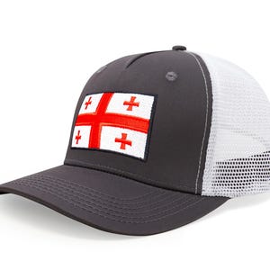 May include: A charcoal gray baseball cap with a white mesh back. The front features a rectangular patch with the flag of Georgia, a red cross on a white background. The cap has a curved brim.