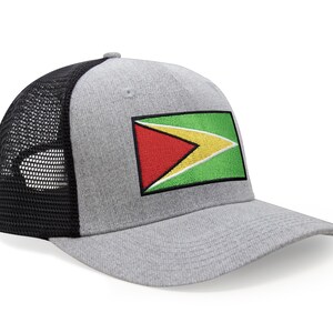 May include: A gray trucker hat with a black mesh back and a curved brim. The front of the hat features an embroidered patch of the Guyana flag, with red, yellow, green, and black colors.