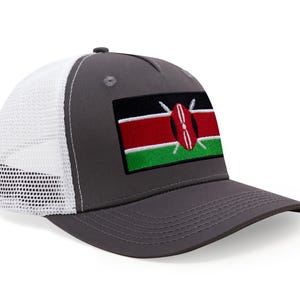May include: A gray trucker hat with a white mesh back and a curved brim. The front panel features an embroidered patch of the Kenyan flag, with black, red, green, and white colors. The hat has a snap closure.