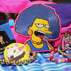 May include: A collection of pop culture themed accessories, including a patch of a cartoon character with blue hair, a pin with a Cher graphic, and a pin with a possum illustration. Also, a pin with a purse graphic and the text "That's My Purse! I Don't Know You!" is visible.