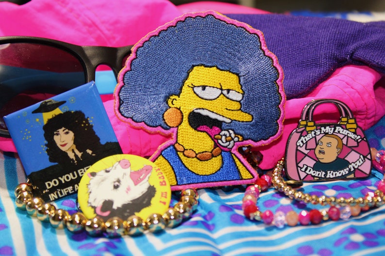 May include: A collection of pop culture themed accessories, including a patch of a cartoon character with blue hair, a pin with a Cher graphic, and a pin with a possum illustration. Also, a pin with a purse graphic and the text "That's My Purse! I Don't Know You!" is visible.