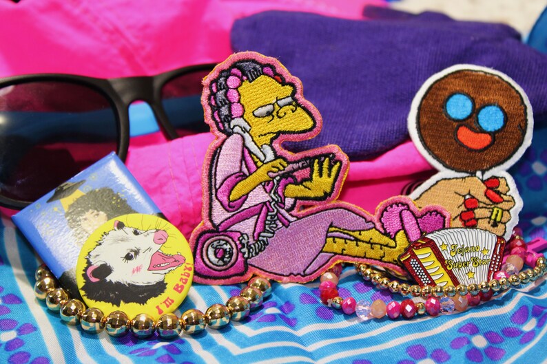 Peut inclure: Une collection d'accessoires comprenant des lunettes de soleil noires, un badge avec un opossum de dessin anim&eacute; et le texte "I'M BABY!", et un &eacute;cusson d'un personnage de dessin anim&eacute;. &Eacute;galement, un bracelet dor&eacute; et un bracelet de perles roses et rouges.