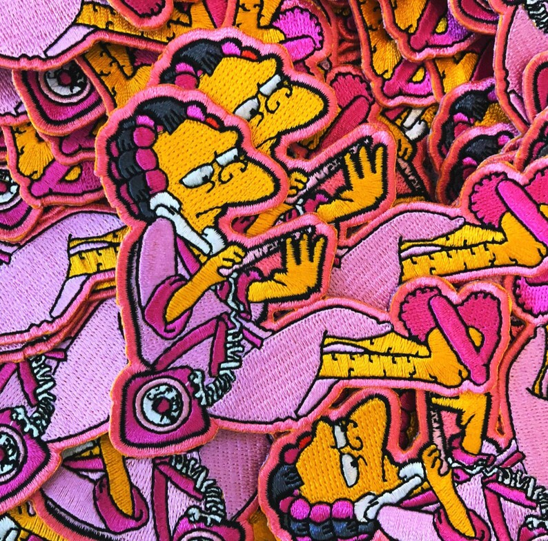 Peut inclure: &Eacute;cusson brod&eacute; repr&eacute;sentant un personnage de dessin anim&eacute; en robe rose, assis et tenant un combin&eacute; t&eacute;l&eacute;phonique. Le personnage a la peau jaune, les cheveux noirs et est soulign&eacute; de rose. L'&eacute;cusson a une bordure rose et est pos&eacute; sur un fond d'&eacute;cussons similaires.