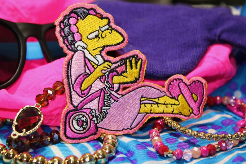 Peut inclure: &Eacute;cusson brod&eacute; repr&eacute;sentant un personnage de dessin anim&eacute; en peignoir rose, bigoudis et pantoufles, tenant un t&eacute;l&eacute;phone. L'&eacute;cusson est entour&eacute; de bijoux, de lunettes de soleil et de tissus color&eacute;s. Le personnage est jaune avec des lunettes.