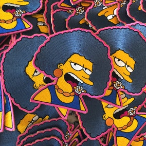 May include: Embroidered patches featuring a cartoon character with blue hair, a yellow face, and a blue dress. The patches have a pink border and a gold necklace. The character is making a surprised face.