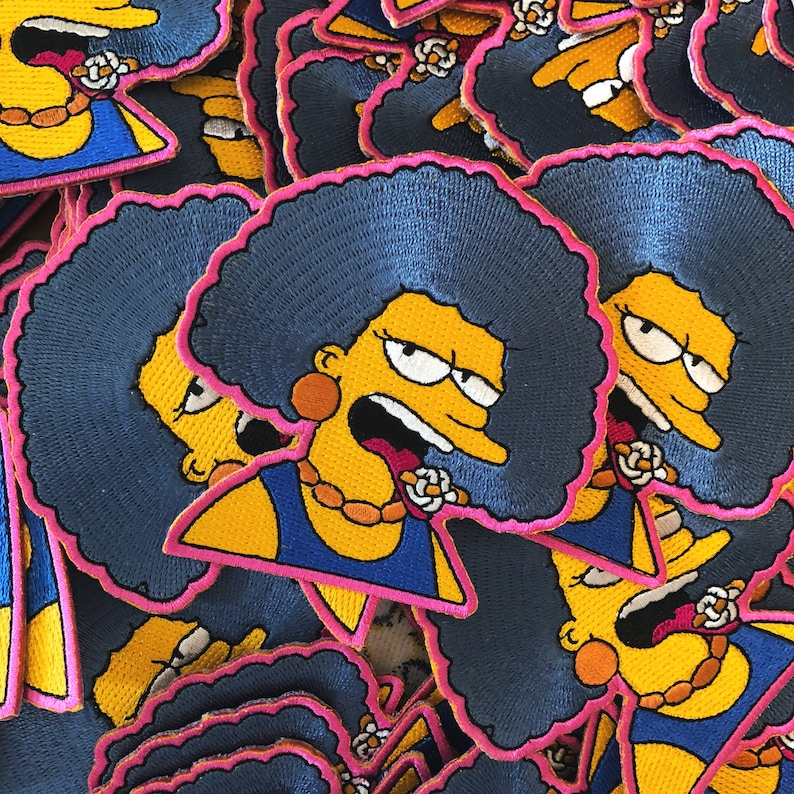 May include: Embroidered patches featuring a cartoon character with blue hair, a yellow face, and a blue dress. The patches have a pink border and a gold necklace. The character is making a surprised face.