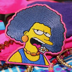 May include: Embroidered patch featuring a cartoon character with yellow skin, blue hair, and a blue dress. The patch is outlined in pink and set against a backdrop of pink and blue fabric, sunglasses, and beaded jewelry.