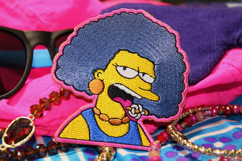May include: Embroidered patch featuring a cartoon character with yellow skin, blue hair, and a blue dress. The patch is outlined in pink and set against a backdrop of pink and blue fabric, sunglasses, and beaded jewelry.