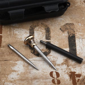 Op de afbeelding: Een meerdelige metalen gereedschapsset bestaande uit een puntige metalen staaf, een centraal handvatonderdeel met een messingkleurige bovenkant met het woord SKULL, en een apart zwart cilindrisch handvatstuk, gerangschikt bij een zwarte opbergdoos.