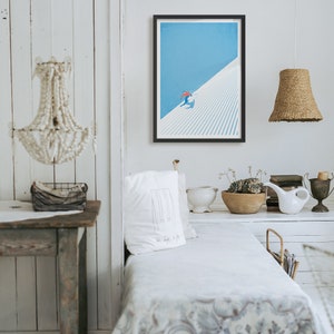 May include: A framed art print depicting a skier on a snow-covered slope against a blue sky. The skier wears a red jacket. The artwork is in a black frame and hangs on a white wall. The room has a minimalist, rustic style.