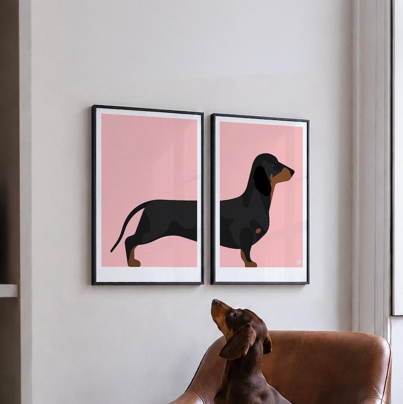 May include: Two framed wall art prints featuring a black and tan dachshund illustration split across two panels. The dog is depicted against a solid light pink background. Each print is enclosed in a thin black frame, creating a cohesive set.