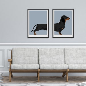 May include: Two framed wall art prints featuring a split illustration of a black and tan dachshund. The left print shows the rear of the dog, while the right print shows the front, both set against a solid light blue background in black frames.