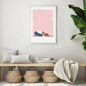 Puede incluir: Una ilustraci&oacute;n enmarcada de una persona haciendo yoga en una postura de ni&ntilde;o sobre un fondo rosa. La ilustraci&oacute;n est&aacute; colgada en una pared blanca sobre un banco de madera con una manta blanca, dos almohadas y tres cestas tejidas.