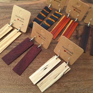 May include: A collection of handmade wooden earrings in various colors and wood grains. The earrings are rectangular and hang from silver-colored hooks. Each pair is displayed on a small, brown card with the text "Handmade with love."
