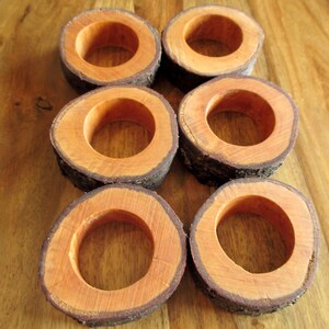 Puede incluir: Seis servilleteros de madera con una est&eacute;tica natural y r&uacute;stica. Cada anillo es una rodaja circular de madera, con un interior marr&oacute;n rojizo c&aacute;lido y un exterior de corteza oscura y texturizada. Los anillos est&aacute;n dispuestos sobre una superficie de madera.