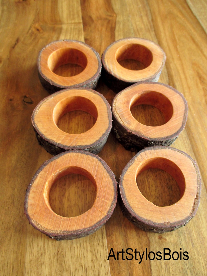 Puede incluir: Seis servilleteros de madera con una est&eacute;tica natural y r&uacute;stica. Cada anillo es una rodaja circular de madera, con un interior marr&oacute;n rojizo c&aacute;lido y un exterior de corteza oscura y texturizada. Los anillos est&aacute;n dispuestos sobre una superficie de madera.