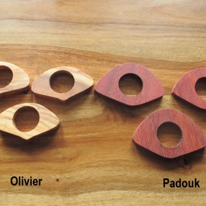 May include: Six wooden napkin rings in two different wood types: olive and padouk. The olive wood rings are light brown, while the padouk rings are a deep reddish-brown. Each ring has a circular opening in the center. The text "Olivier" and "Padouk" are below the rings.