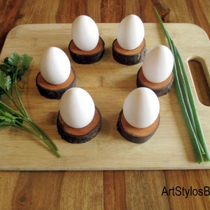 Puede incluir: Seis huevos blancos sobre hueveras individuales de madera, dispuestos en c&iacute;rculo sobre una tabla de cortar de madera. Perejil fresco y cebolletas tambi&eacute;n en la tabla. Las hueveras est&aacute;n hechas de peque&ntilde;as rodajas redondas de madera.