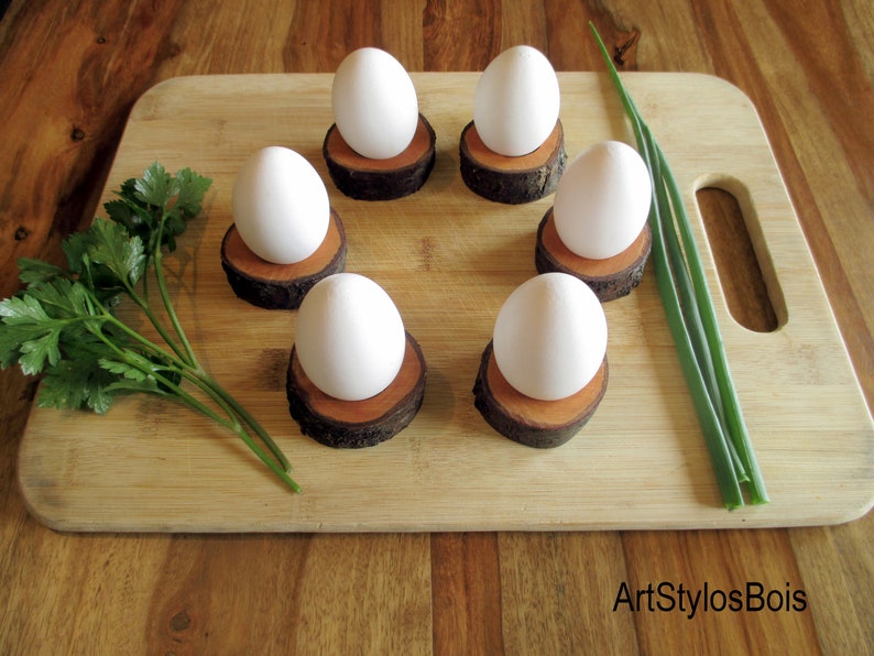 Puede incluir: Seis huevos blancos sobre hueveras individuales de madera, dispuestos en c&iacute;rculo sobre una tabla de cortar de madera. Perejil fresco y cebolletas tambi&eacute;n en la tabla. Las hueveras est&aacute;n hechas de peque&ntilde;as rodajas redondas de madera.
