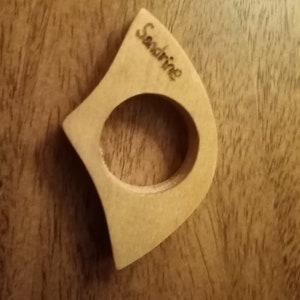 May include: A light-colored wooden ring with a circular opening and a curved, asymmetrical shape. The word "Sandrine" is engraved on the surface. The ring is set against a dark wood grain background.