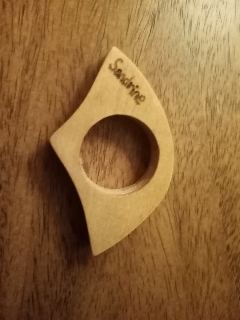 May include: A light-colored wooden ring with a circular opening and a curved, asymmetrical shape. The word "Sandrine" is engraved on the surface. The ring is set against a dark wood grain background.