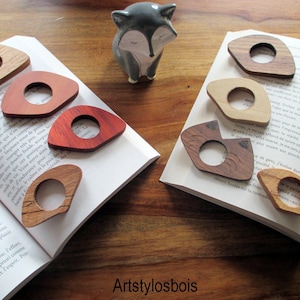 May include: Wooden thumb page holders in various wood grains, including light oak, cherry, and walnut, rest on open books. Each holder has a central hole for the thumb. A ceramic fox figurine is in the background. The shop name "Artstylosbois" is visible.