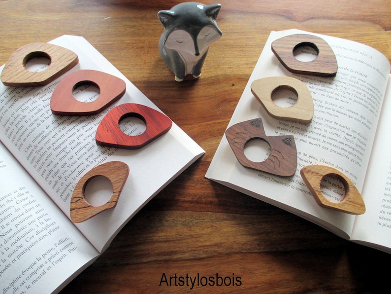 May include: Wooden thumb page holders in various wood grains, including light oak, cherry, and walnut, rest on open books. Each holder has a central hole for the thumb. A ceramic fox figurine is in the background. The shop name "Artstylosbois" is visible.