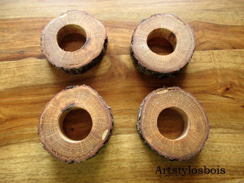 Puede incluir: Cuatro servilleteros de madera hechos de secciones transversales de una rama de &aacute;rbol. Cada anillo es de color marr&oacute;n natural con un agujero central. Los bordes de la corteza son visibles, dispuestos sobre una superficie de madera.