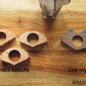 May include: Wooden napkin rings in a variety of designs. Three rings are made of light-colored wood with a circular center and a curved, fan-like shape. One ring is dark wood, shaped like a cat's face, with etched details. The text "H&ecirc;tre &eacute;chauff&eacute;" and "Chat noyer" are visible.