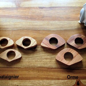 May include: Wooden napkin rings in two wood types, chestnut and elm, are displayed on a wooden surface. The rings have a circular center and a curved, triangular shape. The text "Chataignier" and "Orme" are visible.