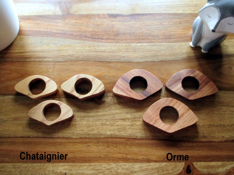 May include: Wooden napkin rings in two wood types, chestnut and elm, are displayed on a wooden surface. The rings have a circular center and a curved, triangular shape. The text "Chataignier" and "Orme" are visible.