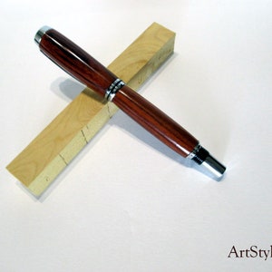 May include: A handcrafted wooden pen with a dark brown finish and silver accents, resting on two light-coloured wooden blocks. The pen's design features a polished, glossy appearance. The pen is a writing instrument.