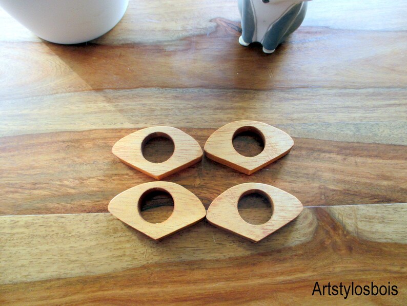 May include: Four wooden napkin rings with a light brown finish. Each ring has an eye-shaped design with a circular opening in the center. The rings are arranged on a wooden surface.