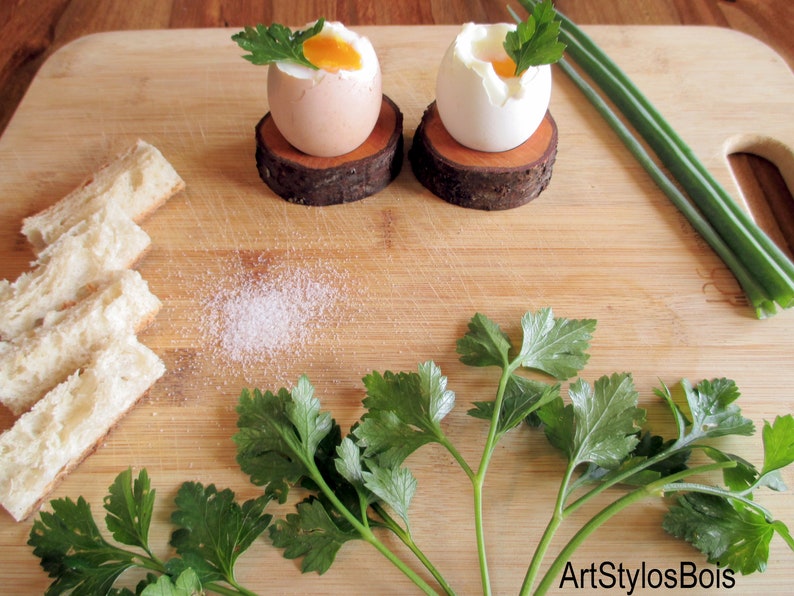 Puede incluir: Dos huevos pasados por agua, uno parcialmente pelado, sobre posavasos de madera r&uacute;stica. Perejil fresco y cebollino se disponen alrededor de los huevos sobre una tabla de cortar de madera, con rebanadas de pan y una pizca de sal.