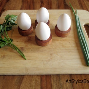 Puede incluir: Cuatro huevos blancos en hueveras de madera sobre una tabla de cortar de bamb&uacute;, con perejil y cebolletas. Las hueveras son peque&ntilde;as, redondas y de madera oscura. La imagen est&aacute; bien iluminada y tiene una est&eacute;tica r&uacute;stica y natural.