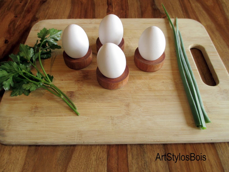 Puede incluir: Cuatro huevos blancos en hueveras de madera sobre una tabla de cortar de bamb&uacute;, con perejil y cebolletas. Las hueveras son peque&ntilde;as, redondas y de madera oscura. La imagen est&aacute; bien iluminada y tiene una est&eacute;tica r&uacute;stica y natural.