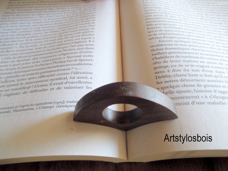 May include: Dark brown wooden book page holder with a circular opening, resting on an open book. The holder has a curved, fan-like shape. The text "Artstylosbois" is visible.