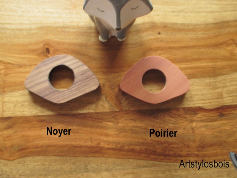 May include: Two wooden, triangular-shaped coasters with a circular hole in the center. One is made of walnut, the other of copper-colored pearwood. The word "Noyer" is printed below the walnut coaster, and "Poirier" is printed below the pearwood coaster.