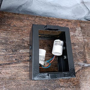 May include: A black, rectangular outdoor light fixture with two white bulb sockets and exposed wiring. The fixture is mounted on a weathered wooden surface. The light fixture is designed to hold two bulbs.