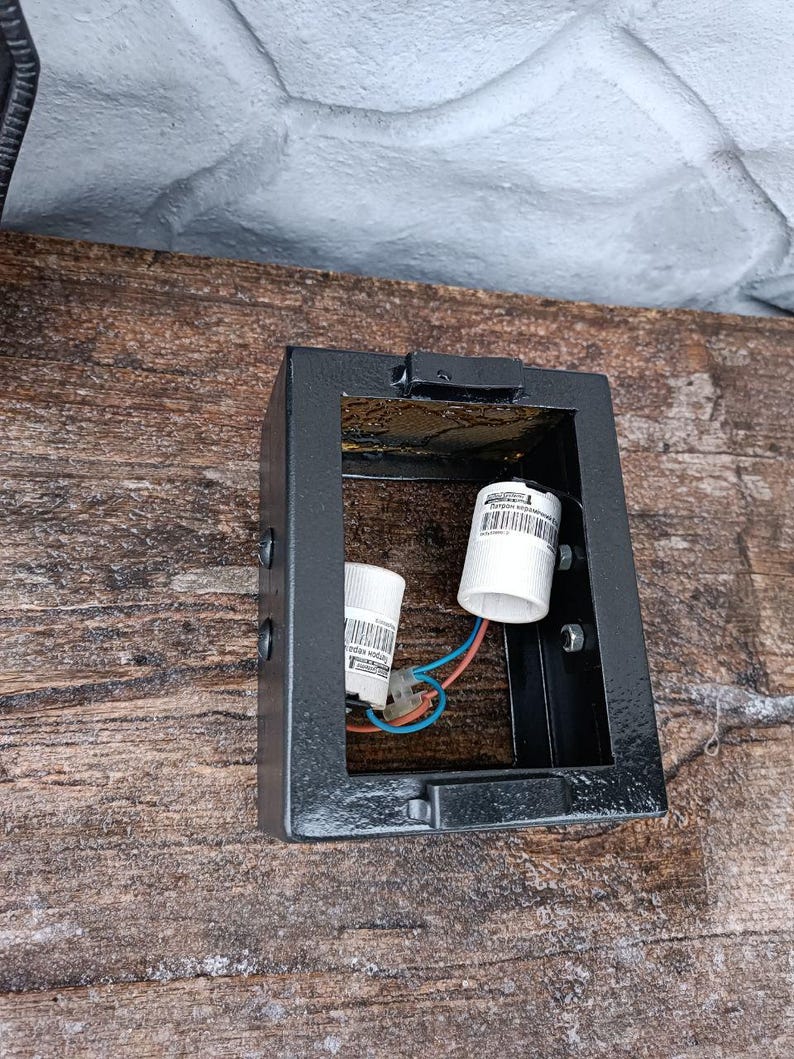 May include: A black, rectangular outdoor light fixture with two white bulb sockets and exposed wiring. The fixture is mounted on a weathered wooden surface. The light fixture is designed to hold two bulbs.
