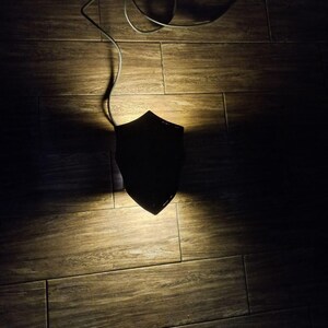 May include: A black, shield-shaped wall sconce with a cord, casting a warm light on a wooden floor. The sconce is the focal point, with the cord extending upwards. The overall aesthetic is minimalist and modern.