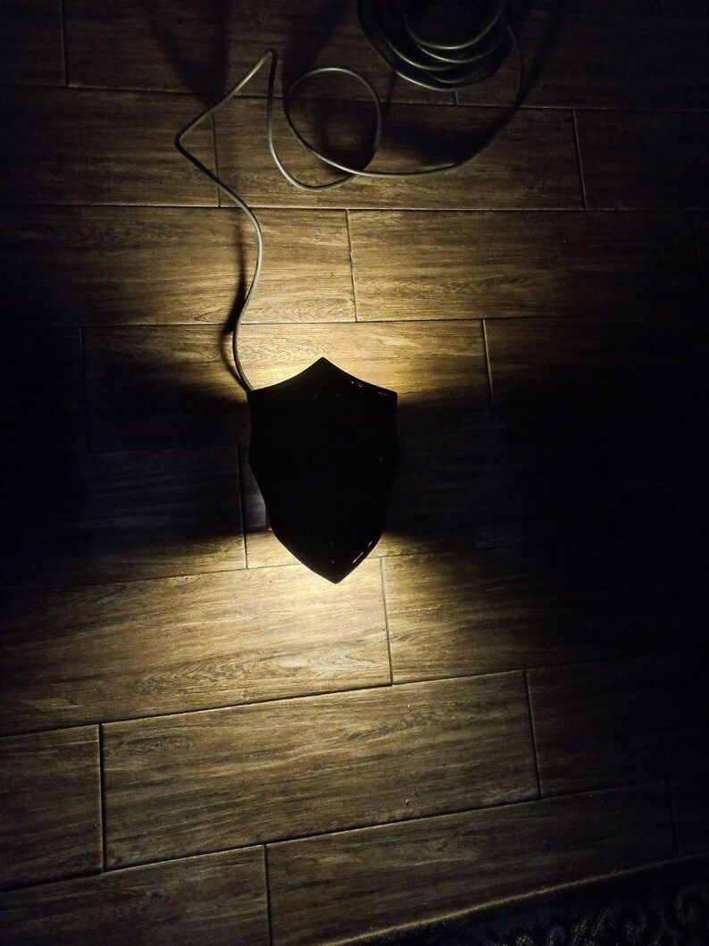 May include: A black, shield-shaped wall sconce with a cord, casting a warm light on a wooden floor. The sconce is the focal point, with the cord extending upwards. The overall aesthetic is minimalist and modern.