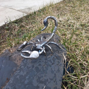 May include: A silver metal sculpture of a scorpion, with a detailed design, sitting on a dark gray rock.