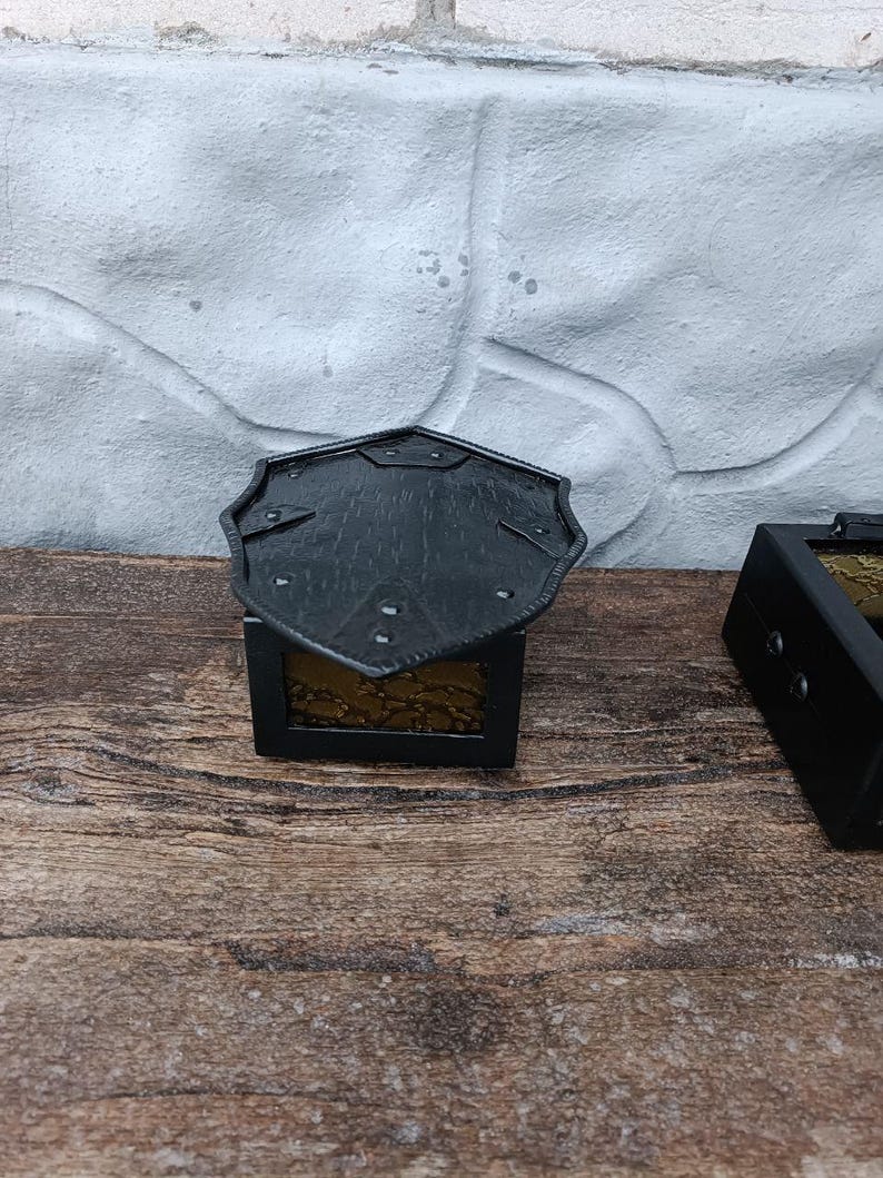 May include: A small, black metal box with a decorative, shield-shaped lid. The box has a rectangular shape with a gold-colored interior. The box is sitting on a wooden surface.