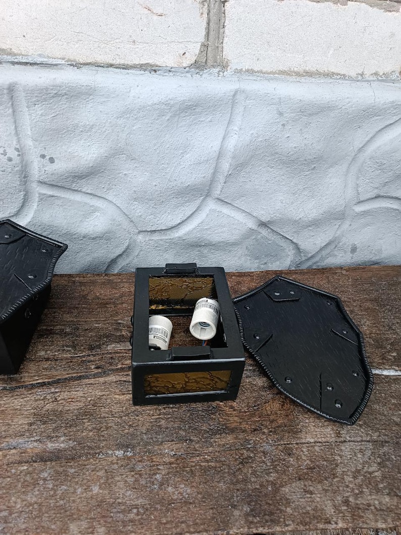 May include: Black metal lantern with amber-colored glass panels. The lantern has a rectangular shape with a hinged door and two light bulb sockets inside. The lantern is sitting on a weathered wooden surface.